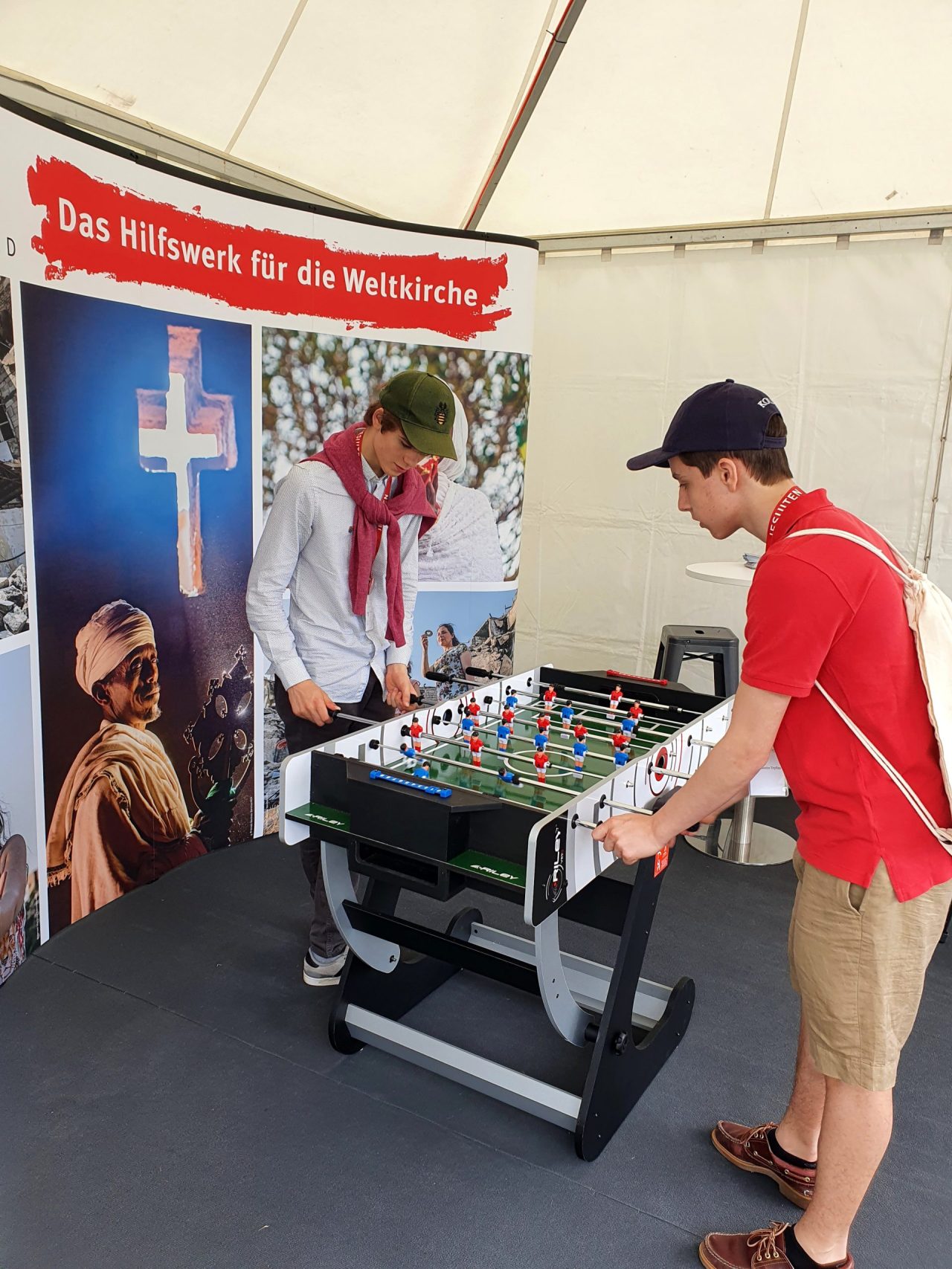 Zwei Jugendliche spielen Tischfußball am Informationsstand von KIRCHE IN NOT. Im Hintergrund informiert ein Roll-up über die Arbeit des Hilfswerks für die Weltkirche.