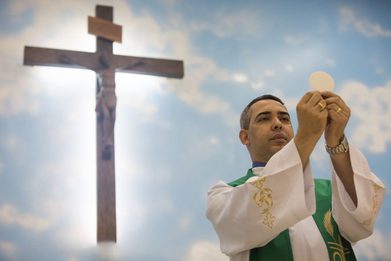 Ein katholischer Priester erhebt während der Eucharistiefeier die Hostie. Im Hintergrund ist ein großes Kruzifix zu sehen.