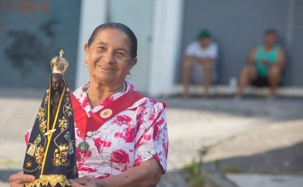Eine Gläubige hält die Statue Unserer Lieben Frau von Aparecida, der Patronin Brasiliens, in den Händen. Sie lächelt in die Kamera und trägt ein rotes Schulterband.