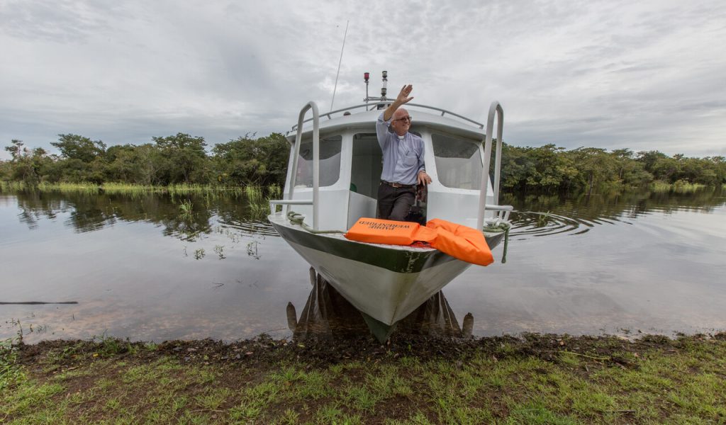 Ein katholischer Priester sitzt in einem Boot am Ufer eines Flusses und winkt. Mit dem Missionsboot erreicht die Kirche Menschen in entlegenen Gebieten.