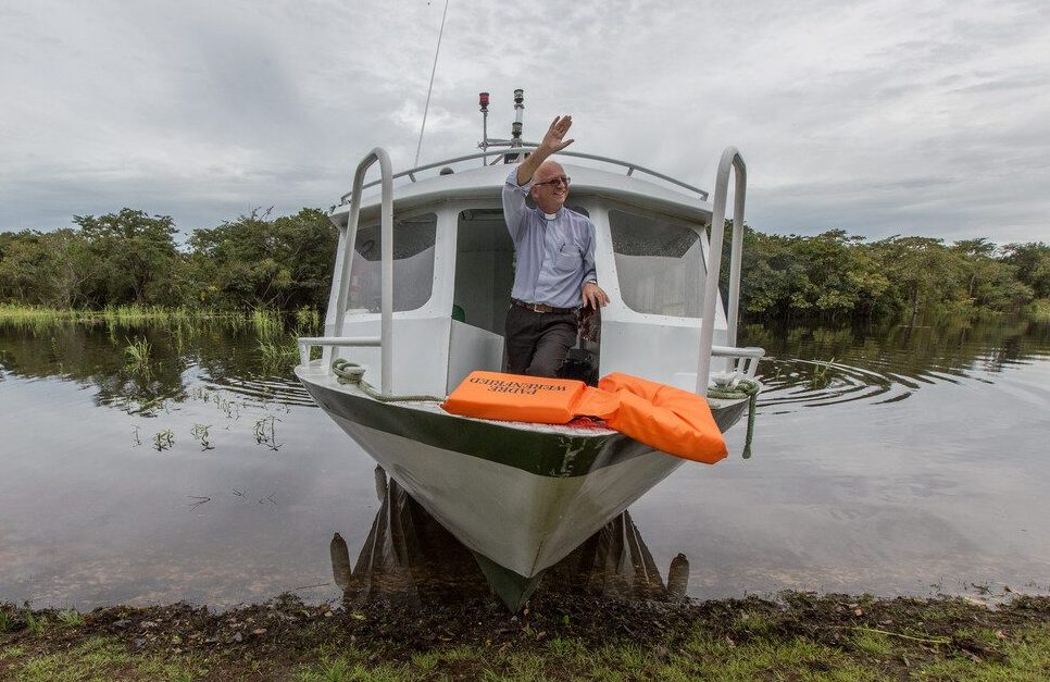 Ein katholischer Priester sitzt in einem Boot am Ufer eines Flusses und winkt. Mit dem Missionsboot erreicht die Kirche Menschen in entlegenen Gebieten.