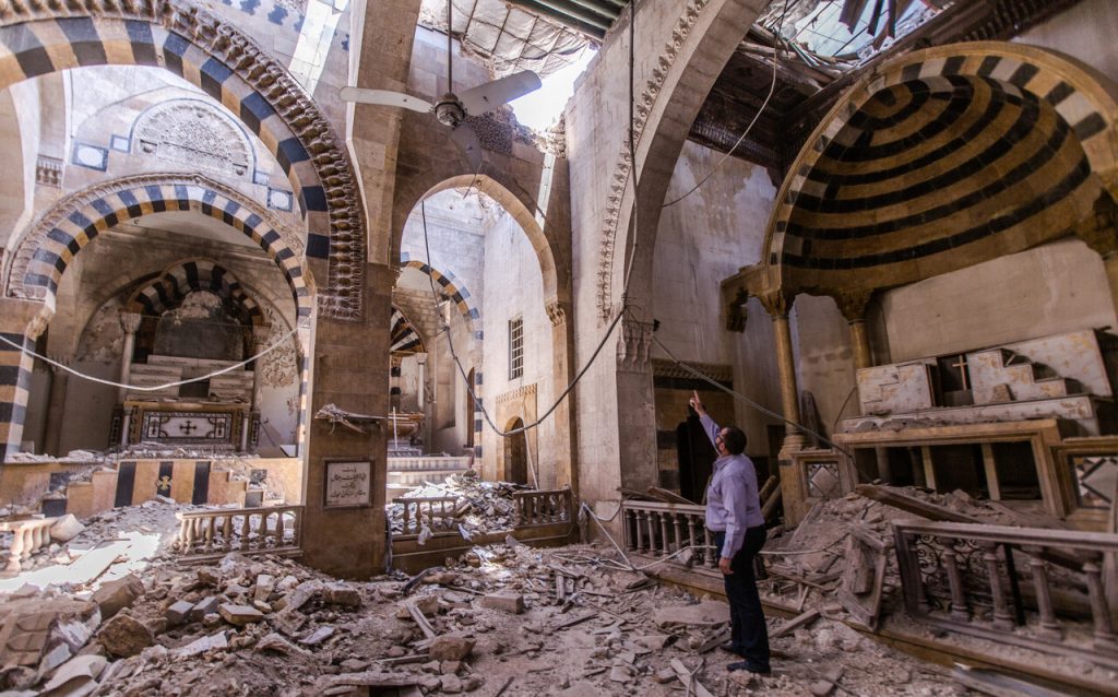 Das Innere einer schwer beschädigten Kirche mit eingestürztem Dach und Trümmern auf dem Boden. Ein Mann steht zwischen den Ruinen und blickt nach oben.