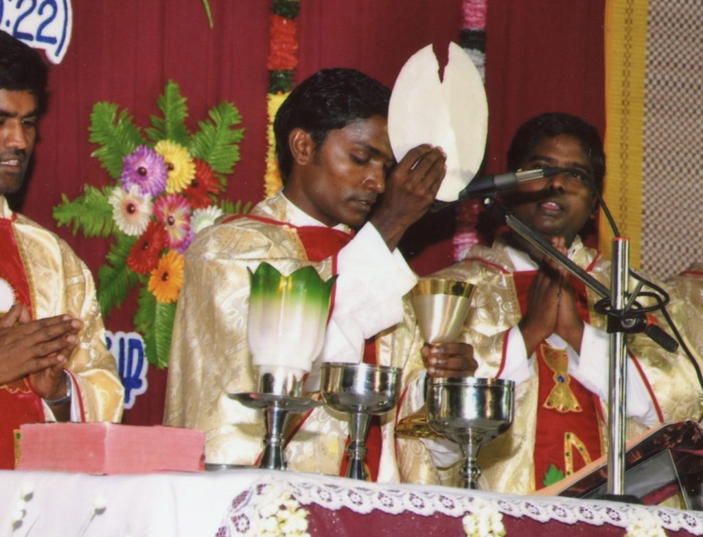 Mehrere katholische Priester feiern gemeinsam die Heilige Messe. Während der Eucharistiefeier erhebt ein Priester die Hostie am Altar, die anderen beten mit gefalteten Händen.