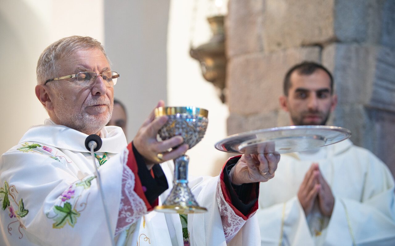 Ein Priester feiert die Heilige Messe und erhebt während der Eucharistiefeier Kelch und Patene. Im Hintergrund steht ein weiterer liturgischer Diener im Gebet.