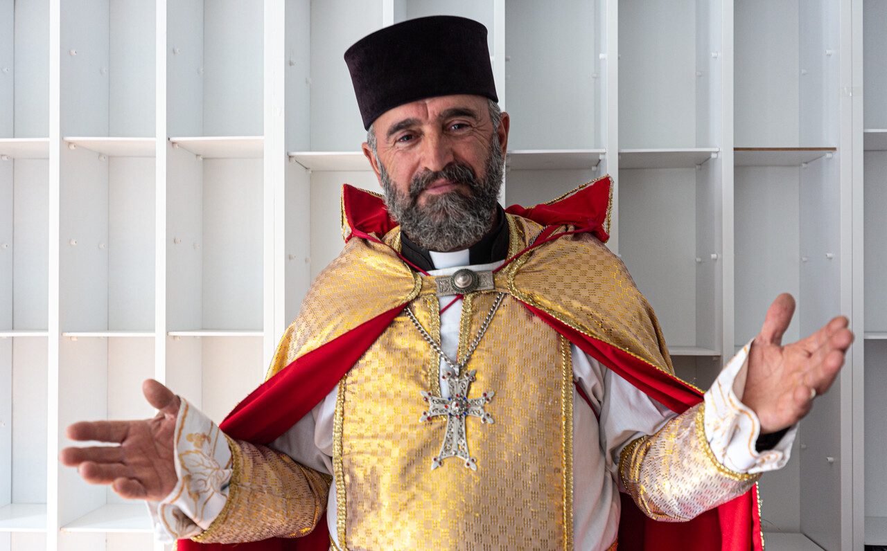 Ein Geistlicher der Ostkirche in liturgischen Gewändern steht mit ausgebreiteten Armen vor einer Wand mit leeren Regalfächern.
