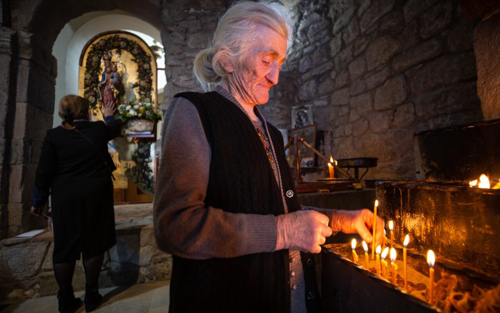 Eine ältere Frau entzündet Kerzen in einer Kirche; im Hintergrund betet eine weitere Frau vor einer Marienstatue.