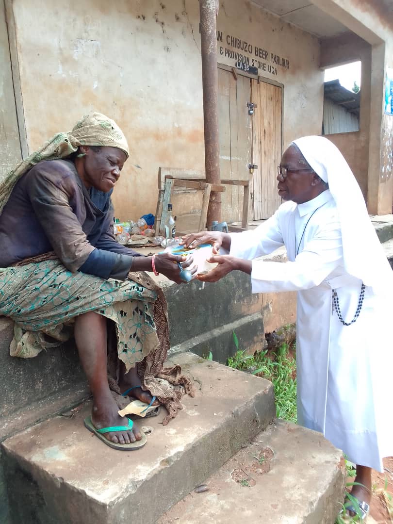 Eine Ordensschwester überreicht einer bedürftigen Frau vor einem Gebäude in Nigeria Lebensmittel. Die Frau sitzt auf einer Treppe und nimmt die Hilfe entgegen.