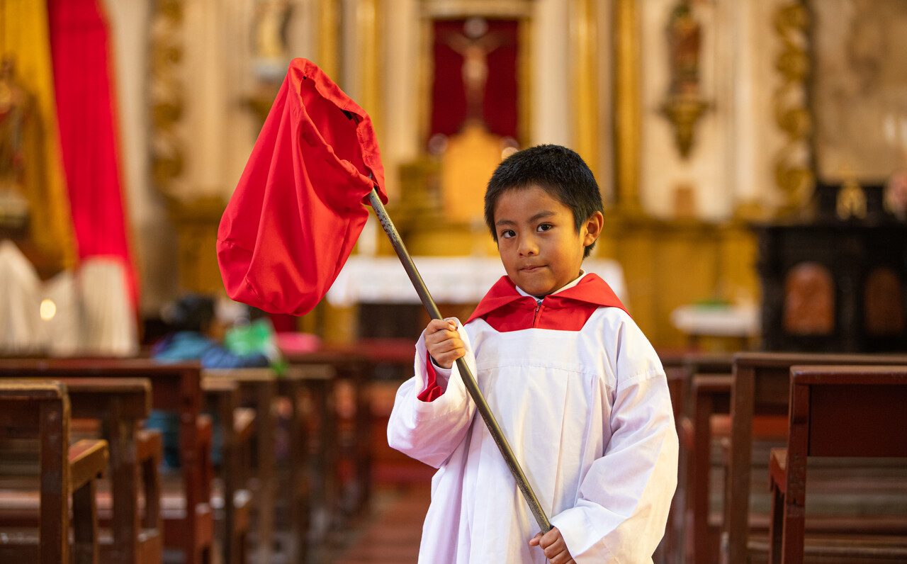 Ein Ministrant in weiß-rotem Gewand steht in einer Kirche und hält einen Kollektenkorb an einer langen Stange. Im Hintergrund ist der Altar zu sehen.