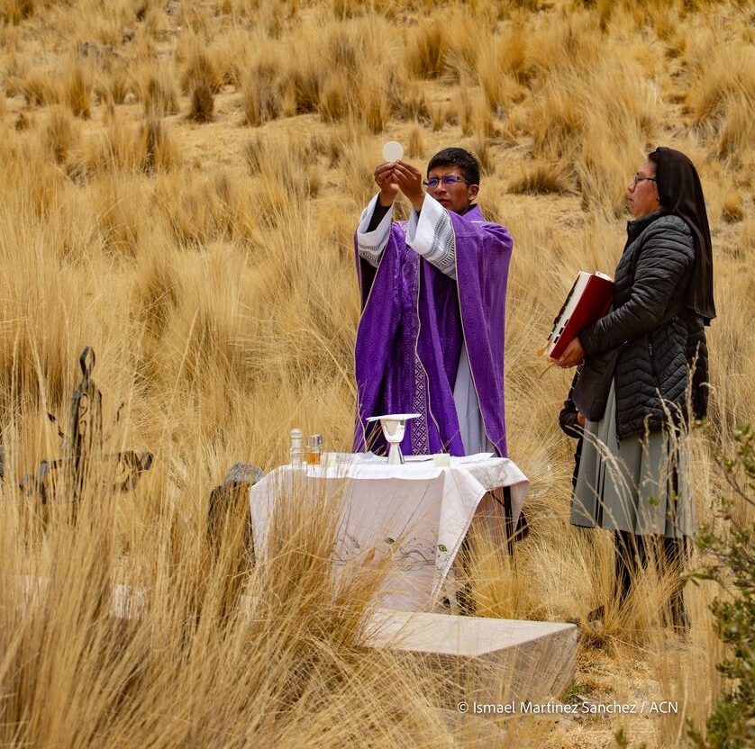 Ein Priester feiert unter freiem Himmel die Heilige Messe und erhebt die Hostie. Neben ihm steht eine Ordensschwester mit einem Messbuch. Die Feier findet in einer ländlichen Hochebene statt.