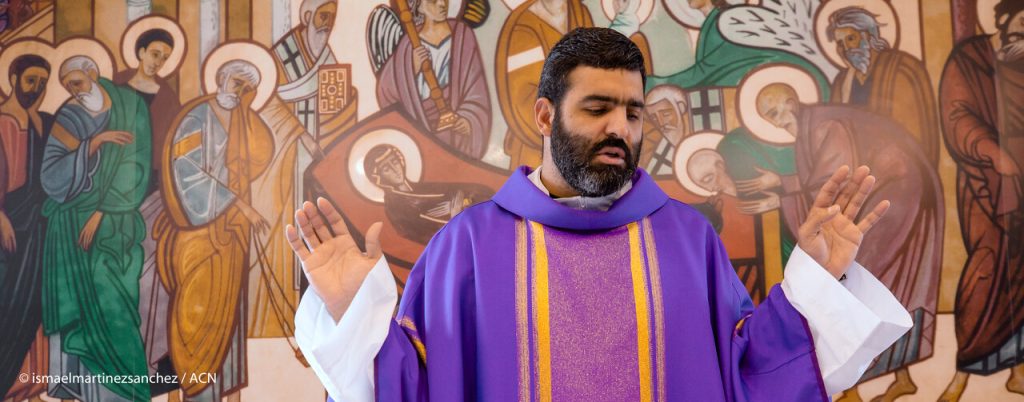 Ein katholischer Priester in violetten liturgischen Gewändern betet mit erhobenen Händen vor einem Wandgemälde mit Heiligendarstellungen.
