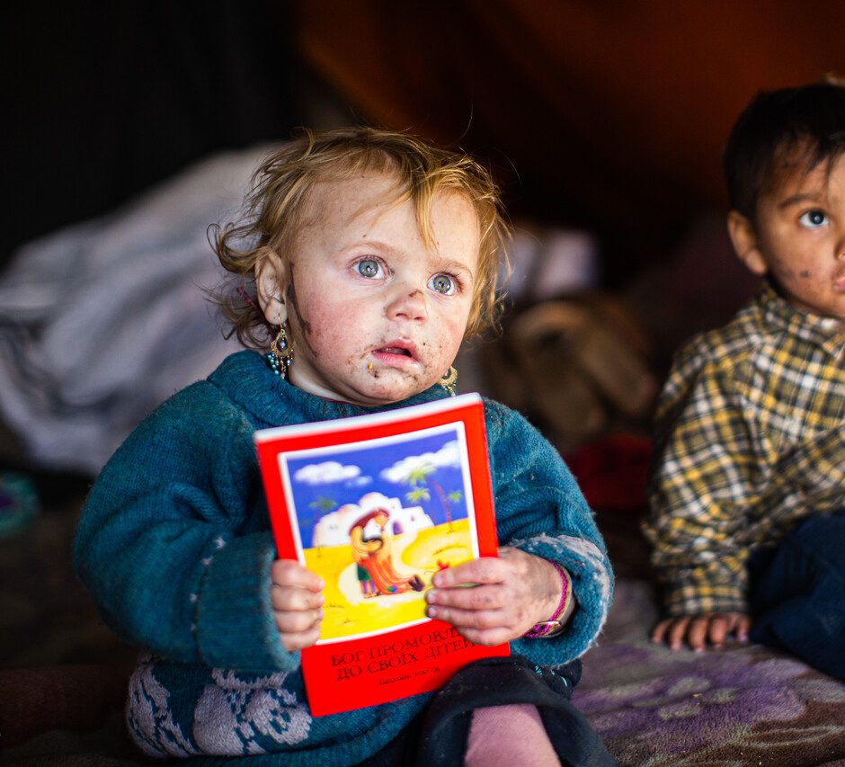 Zwei kleine Kinder sitzen in einer einfachen Unterkunft. Eines der Kinder hält eine Kinderbibel in den Händen und blickt in die Kamera.