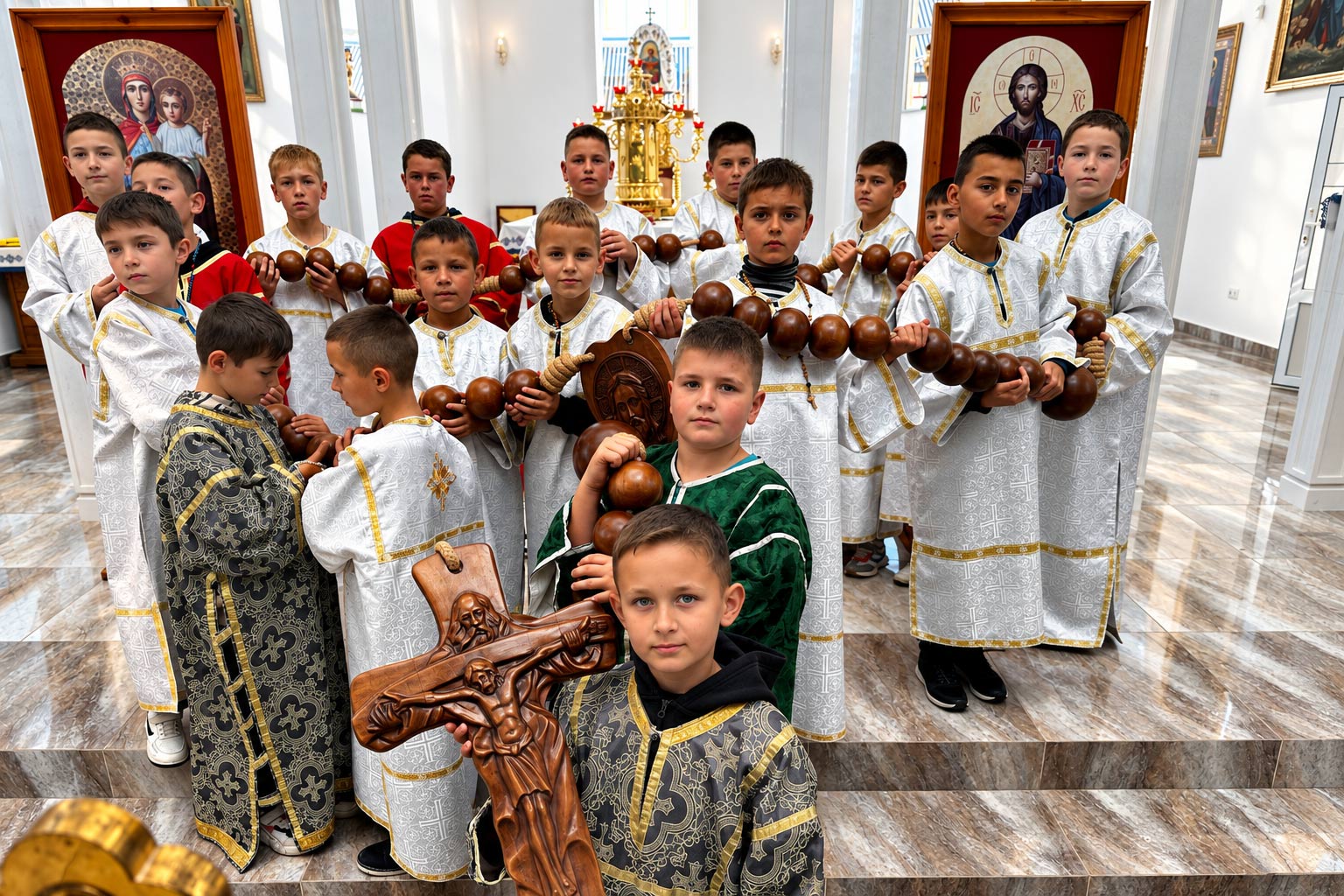 Eine Gruppe von Jungen in liturgischen Gewändern steht in einer Kirche. Mehrere halten einen großen Rosenkranz aus Holzkugeln; im Vordergrund trägt ein Junge ein geschnitztes Holzkreuz.