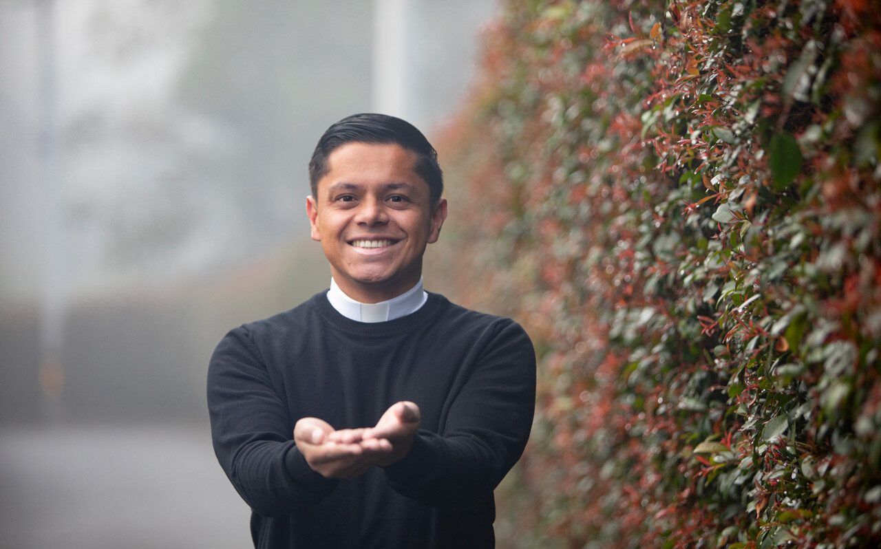 Ein lächelnder junger Priester steht im Freien und hält die Hände einladend nach vorne. Im Hintergrund ist eine Hecke zu sehen.