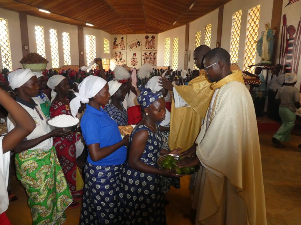 Gläubige bringen während eines Gottesdienstes Gaben zum Altar. Zwei Priester segnen die Menschen und ihre Opfergaben in einer voll besetzten Kirche.