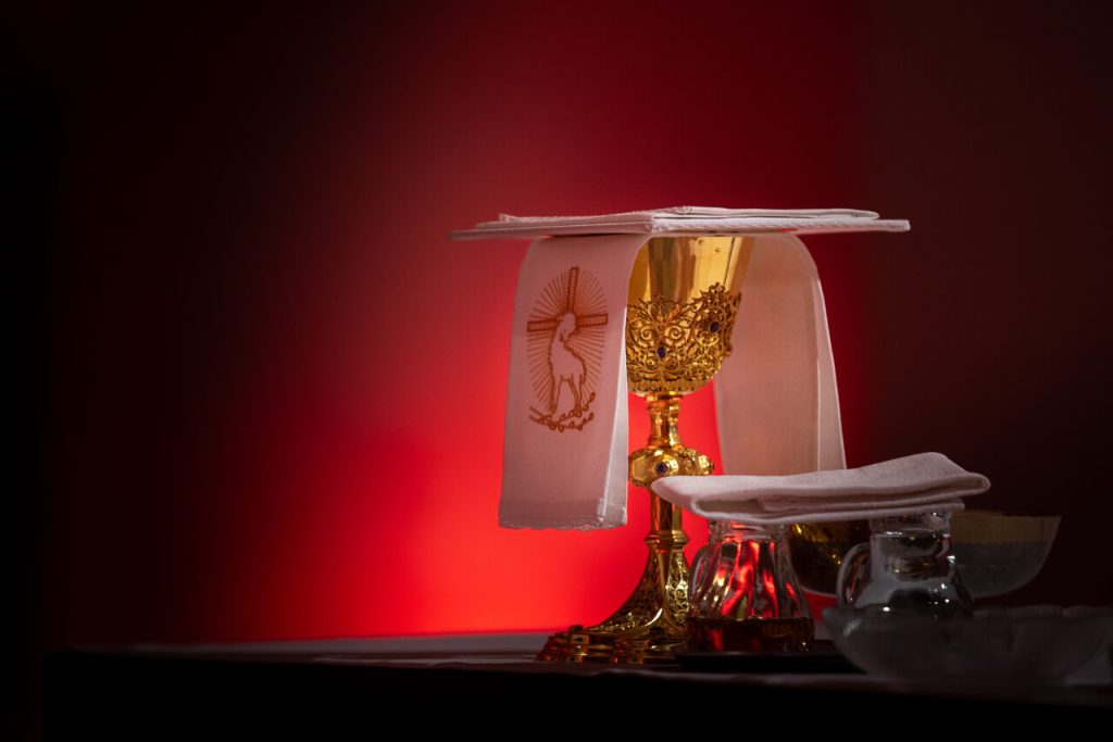 Ein goldener Messkelch mit Palla und Kelchtuch steht auf einem Altar. Daneben befinden sich die Messkännchen für Wein und Wasser vor einem rot beleuchteten Hintergrund
