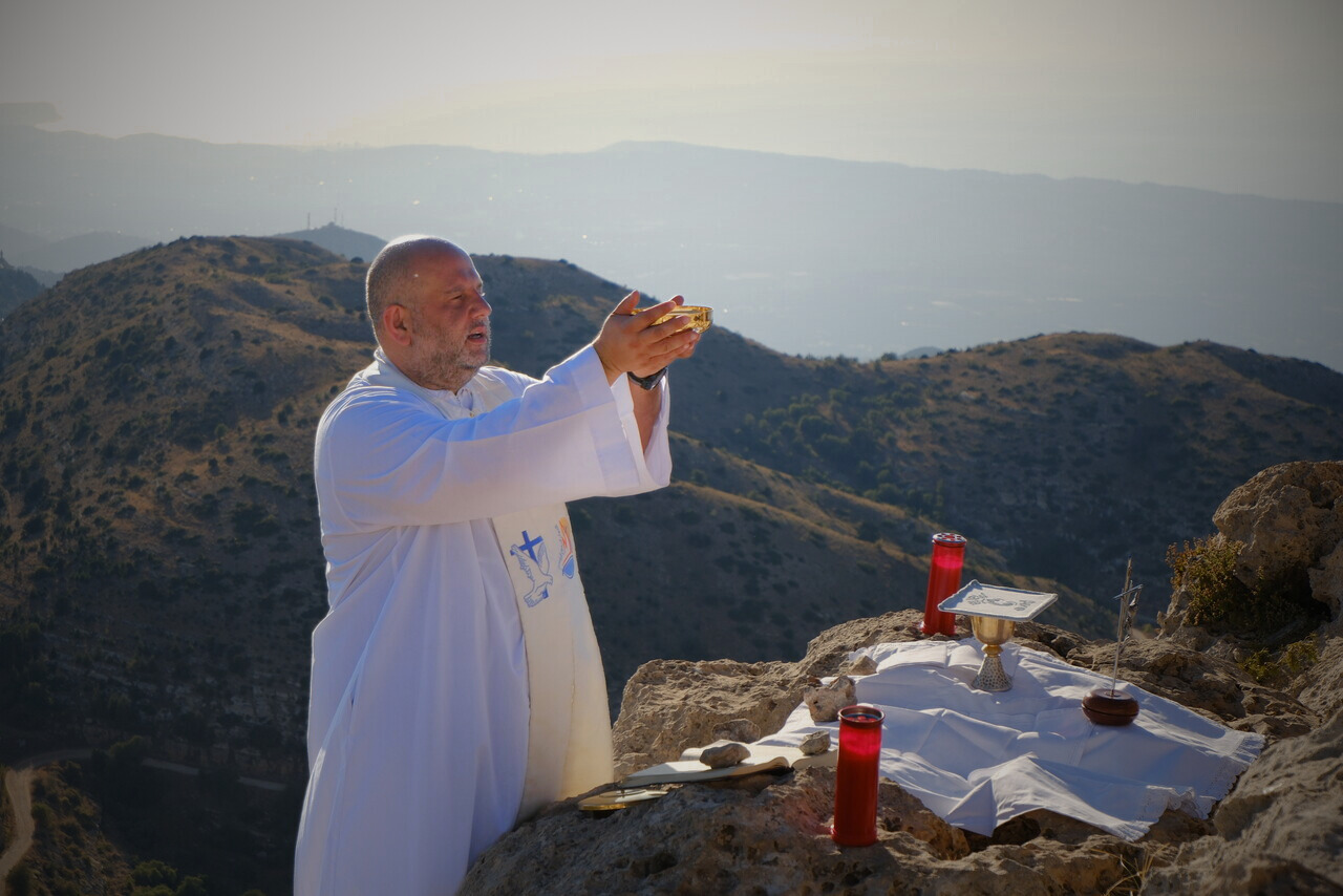 Ein katholischer Priester feiert unter freiem Himmel die Heilige Messe auf einem Berg und erhebt den Kelch. Ein provisorischer Altar mit liturgischen Gegenständen steht auf den Felsen vor einer weiten Berglandschaft