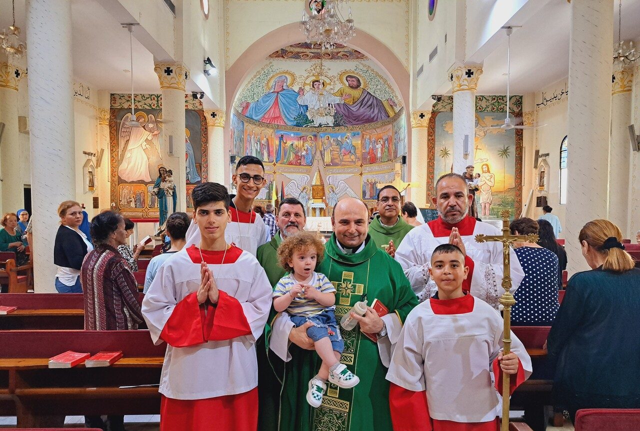 Pater Gabriel Romanelli posiert nach der Heiligen Messe mit Ministranten, Gemeindemitgliedern und einem Kleinkind für ein Gruppenfoto vor dem Altar einer Kirche.