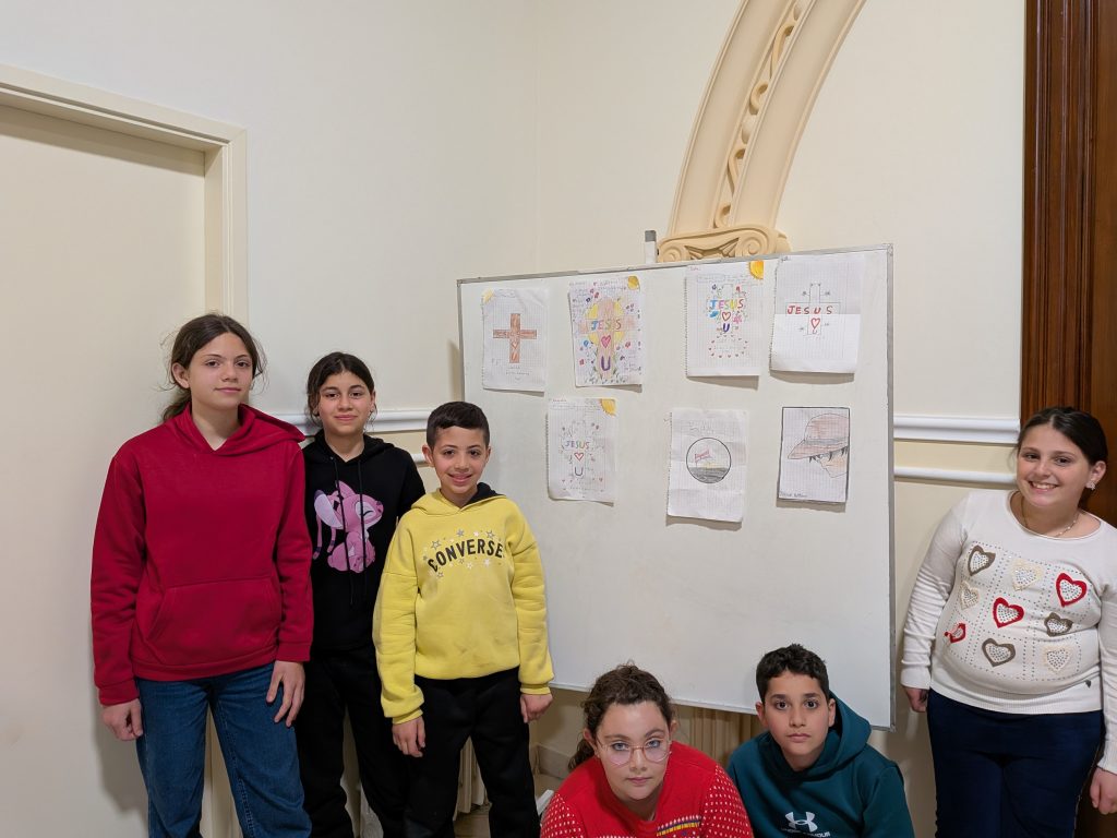 Eine Gruppe von Kindern präsentiert selbstgemalte Bilder mit christlichen Motiven auf einer Pinnwand.