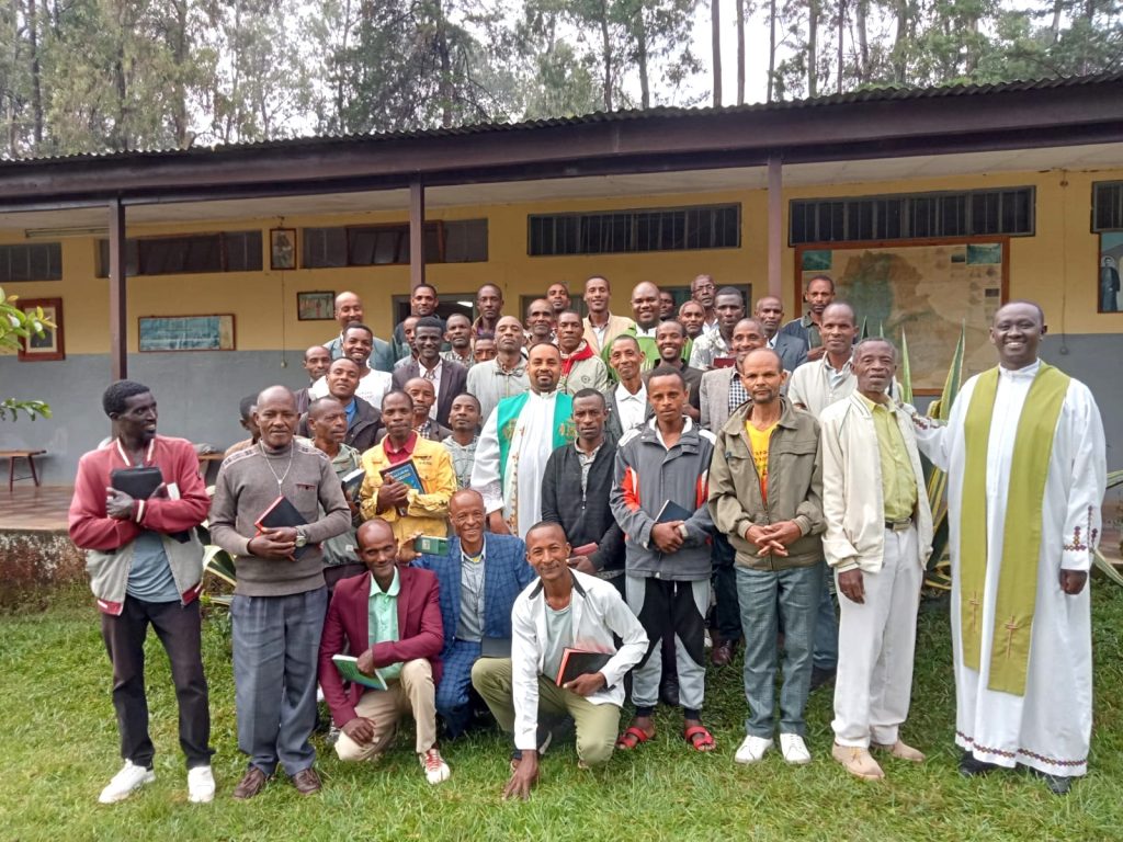 Eine Gruppe von Katecheten und Priestern versammelt sich vor einem kirchlichen Gebäude zu einem Gruppenfoto. Viele halten Bibeln oder religiöse Bücher in den Händen.