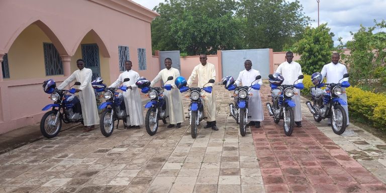 Mehrere Priester stehen mit neuen Motorrädern vor einer kirchlichen Einrichtung. Die Fahrzeuge unterstützen ihre Seelsorge in abgelegenen Gemeinden.
