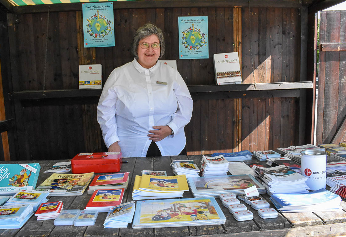Eine Mitarbeiterin von KIRCHE IN NOT steht an einem Informationsstand mit Broschüren, Kinderbibeln und weiteren Materialien. Auf dem Tisch liegen Informationshefte und Spendenmaterialien des Hilfswerks.