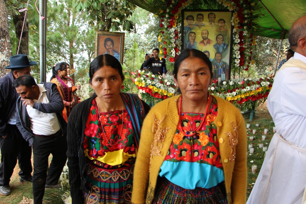 Zwei Frauen in traditioneller indigener Kleidung stehen vor einem Gedenkaltar mit Blumen und Porträts katholischer Märtyrer. Im Hintergrund versammeln sich Gläubige zu einer Gedenkfeier.