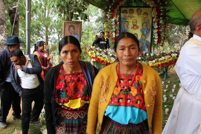 Zwei Frauen in traditioneller indigener Kleidung stehen vor einem Gedenkaltar mit Blumen und Porträts katholischer Märtyrer. Im Hintergrund versammeln sich Gläubige zu einer Gedenkfeier.
