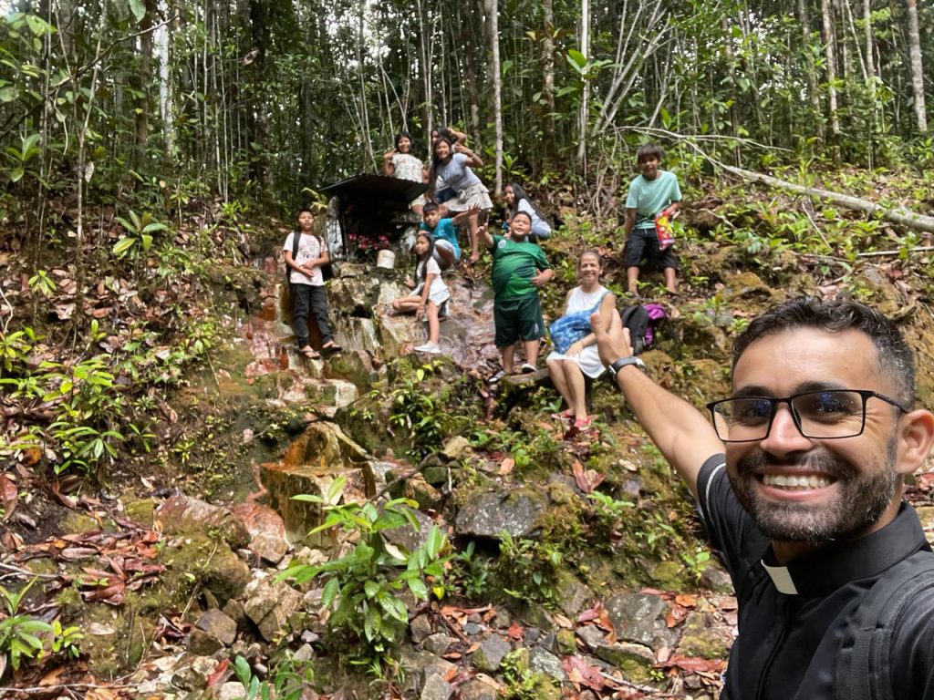 Ein Priester macht ein Selfie mit einer Gruppe von Kindern und Erwachsenen bei einem Ausflug im Regenwald. Die Gruppe steht an einem kleinen Bach zwischen Felsen und dichtem Wald.