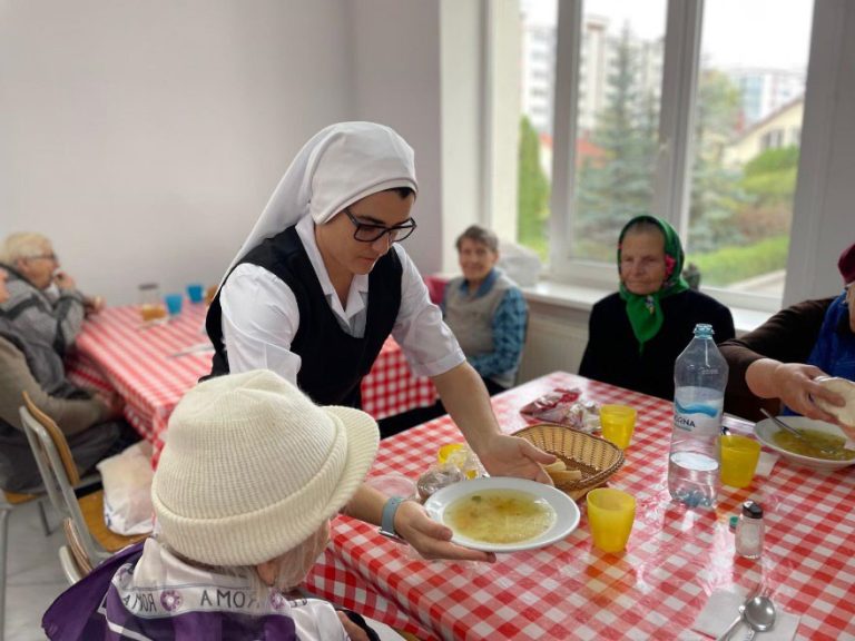 Eine Ordensschwester serviert älteren Menschen in einem Speisesaal eine warme Mahlzeit. Die Seniorinnen sitzen gemeinsam an einem gedeckten Tisch.