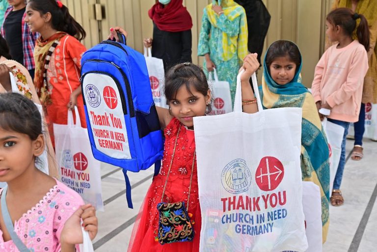 Mehrere Kinder halten Schulrucksäcke und Tragetaschen mit dem Logo von KIRCHE IN NOT in den Händen. Die Hilfspakete wurden im Rahmen einer kirchlichen Unterstützungsaktion verteilt.
