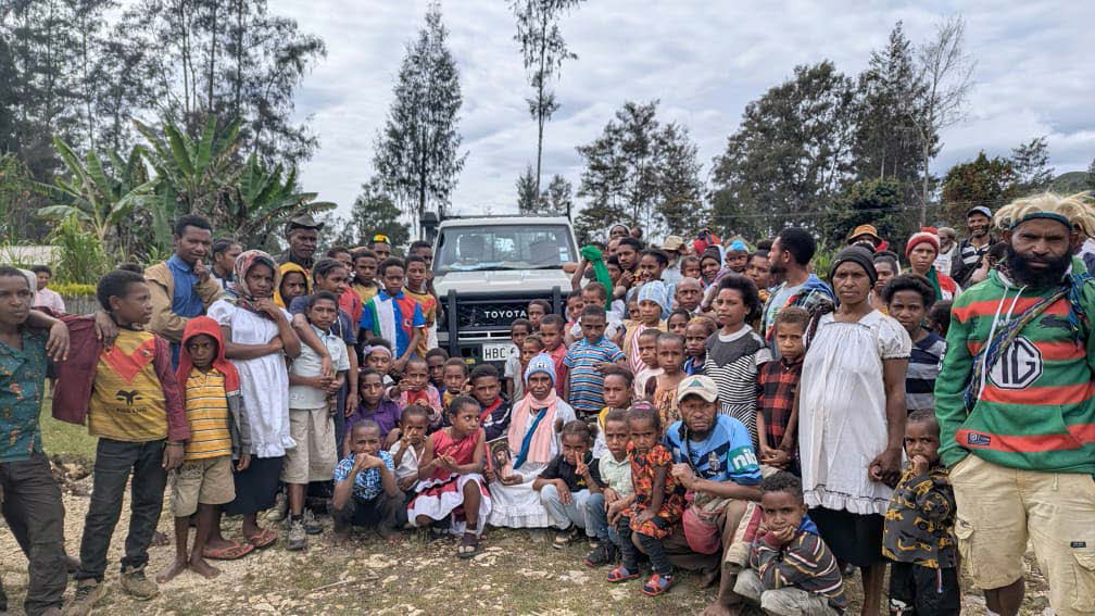 Eine große Gruppe von Erwachsenen und Kindern versammelt sich vor einem Geländewagen in einem Dorf. Die Menschen stehen gemeinsam für ein Gruppenfoto im Freien.