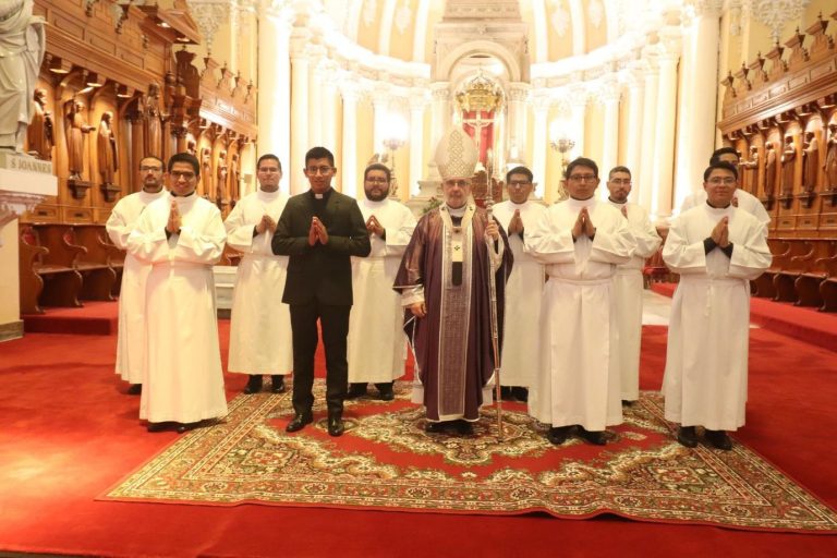 Ein Bischof steht gemeinsam mit Seminaristen und Priestern vor dem Altar einer Kirche. Die Seminaristen tragen liturgische Gewänder und haben die Hände zum Gebet gefaltet.