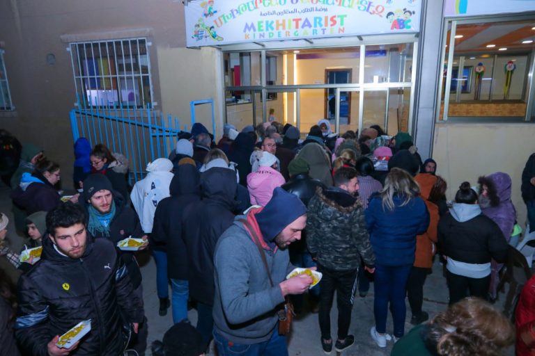 Zahlreiche Menschen warten vor einem Kindergarten auf die Ausgabe von Lebensmitteln. Viele tragen warme Kleidung und halten bereits Mahlzeiten in den Händen.